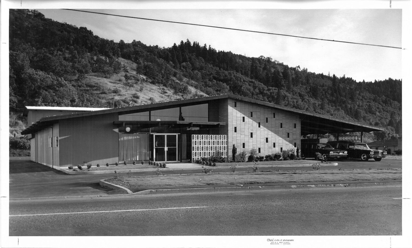 Black-and-white photo of a modern roadside building with parked cars and a wooded hillside behind it