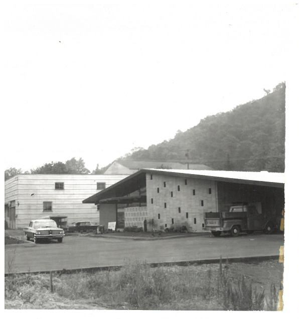 Black-and-white photo of a roadside building with two parked cars and a wooded hillside behind it