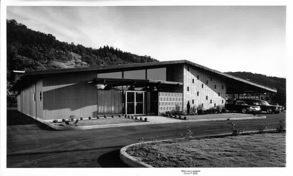 Black-and-white photo of a modern building with a sloped roof and landscaped entrance, set against hills.