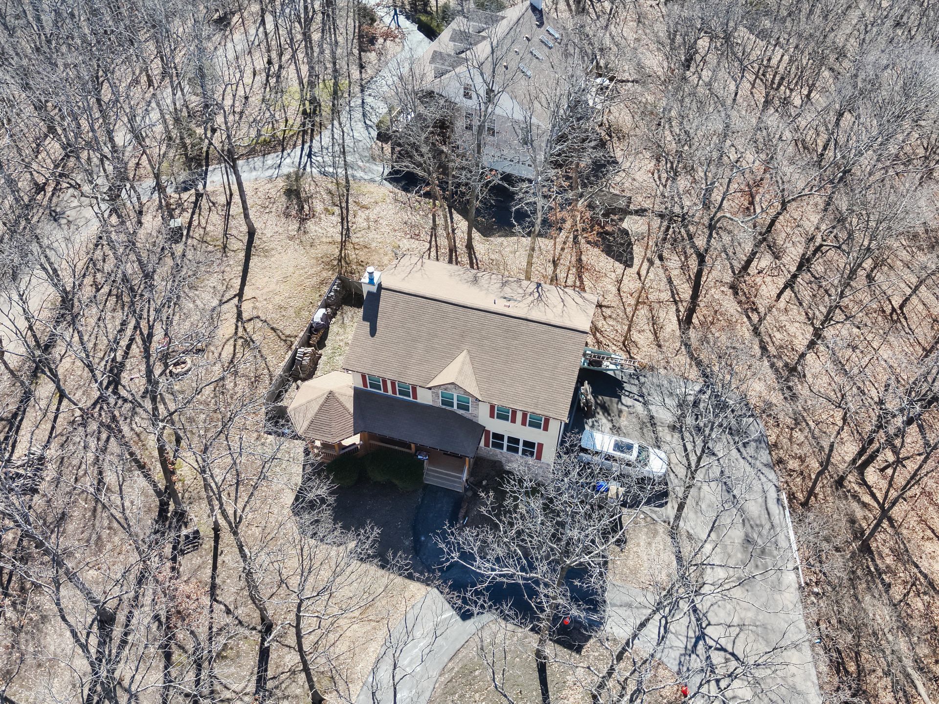 Aerial view of a house surrounded by bare trees; brown roof, white siding, driveway.