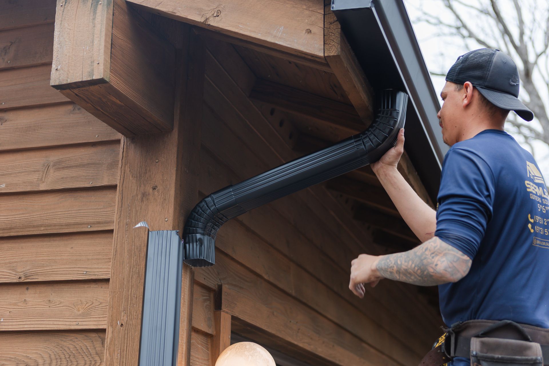 Man in blue shirt installing black gutter on brown wooden building.