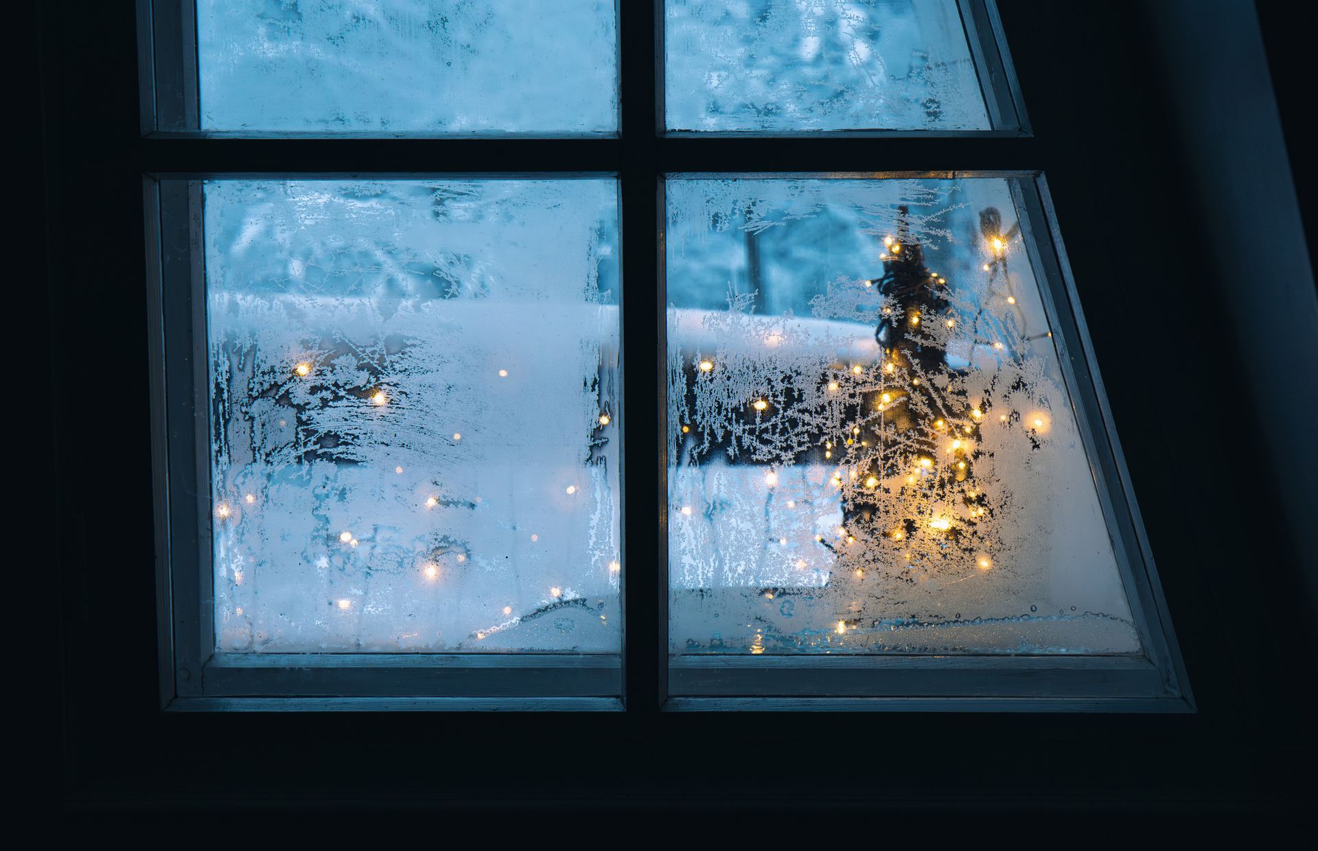 Frosted window with Christmas tree inside, illuminated by warm fairy lights against a cold, blue backdrop.
