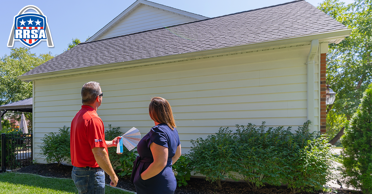 A man in a red shirt and a woman in a blue dress look at papers next to a house with a dark roof.