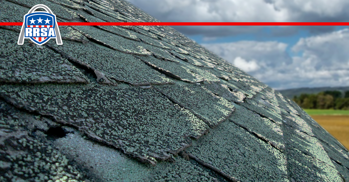 Damaged, weathered shingles on a roof with a logo in the upper left. Sky and fields in the background.