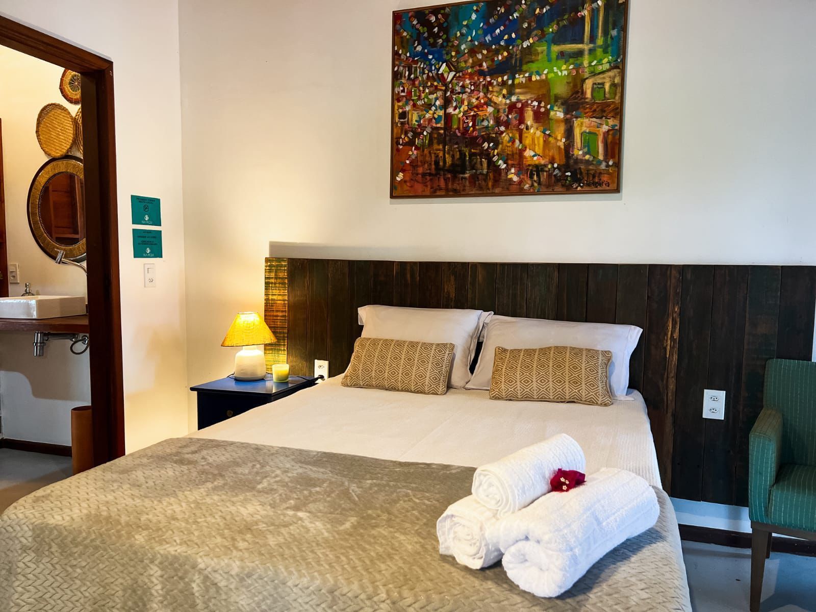 Quarto de hotel com cama arrumada, abajur, obras de arte coloridas na parede e banheiro visível pela porta.