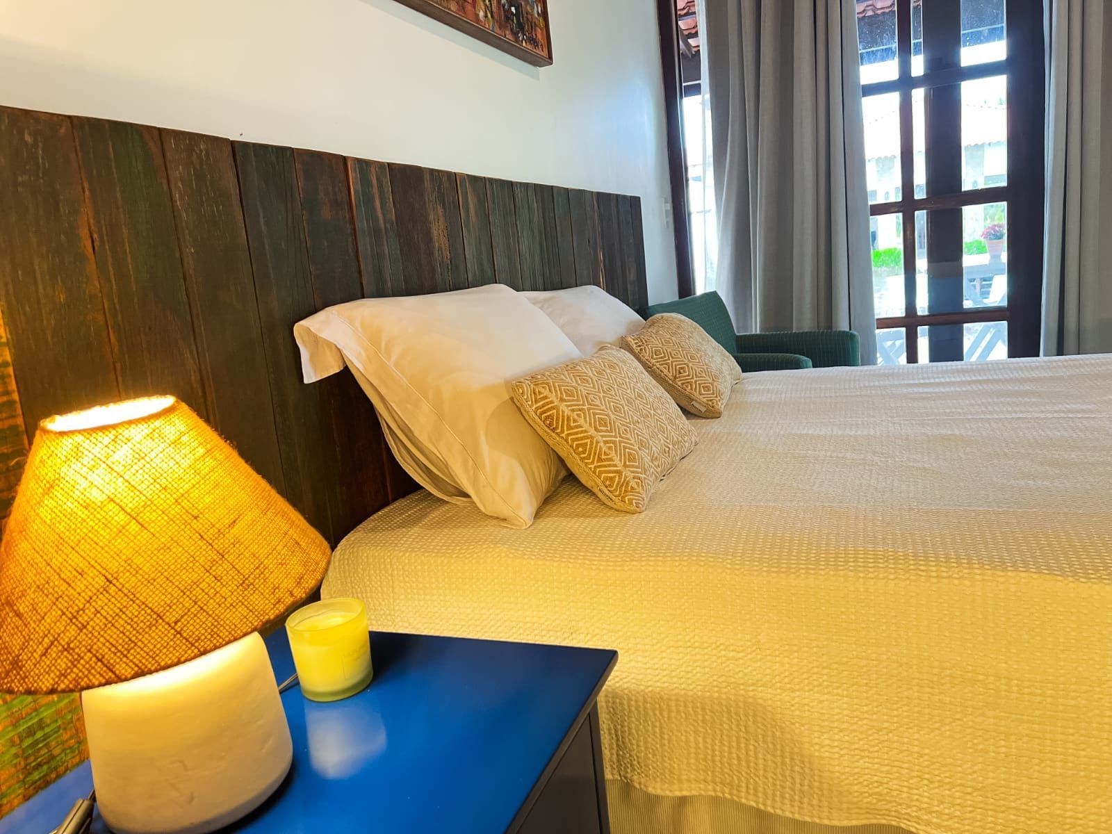 Quarto aconchegante com cama, mesa de cabeceira azul, abajur com luz quente e luz solar entrando pelas janelas altas.