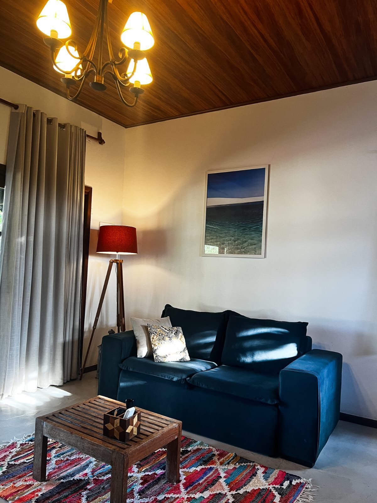 Sala de estar aconchegante com sofá preto, luminária de chão vermelha, mesa de centro, cortinas e teto com painéis de madeira.