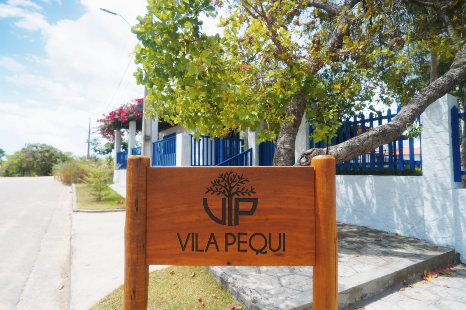 Placa de madeira VIP Vila Pequi ao lado de um prédio azul e branco e uma estrada arborizada.