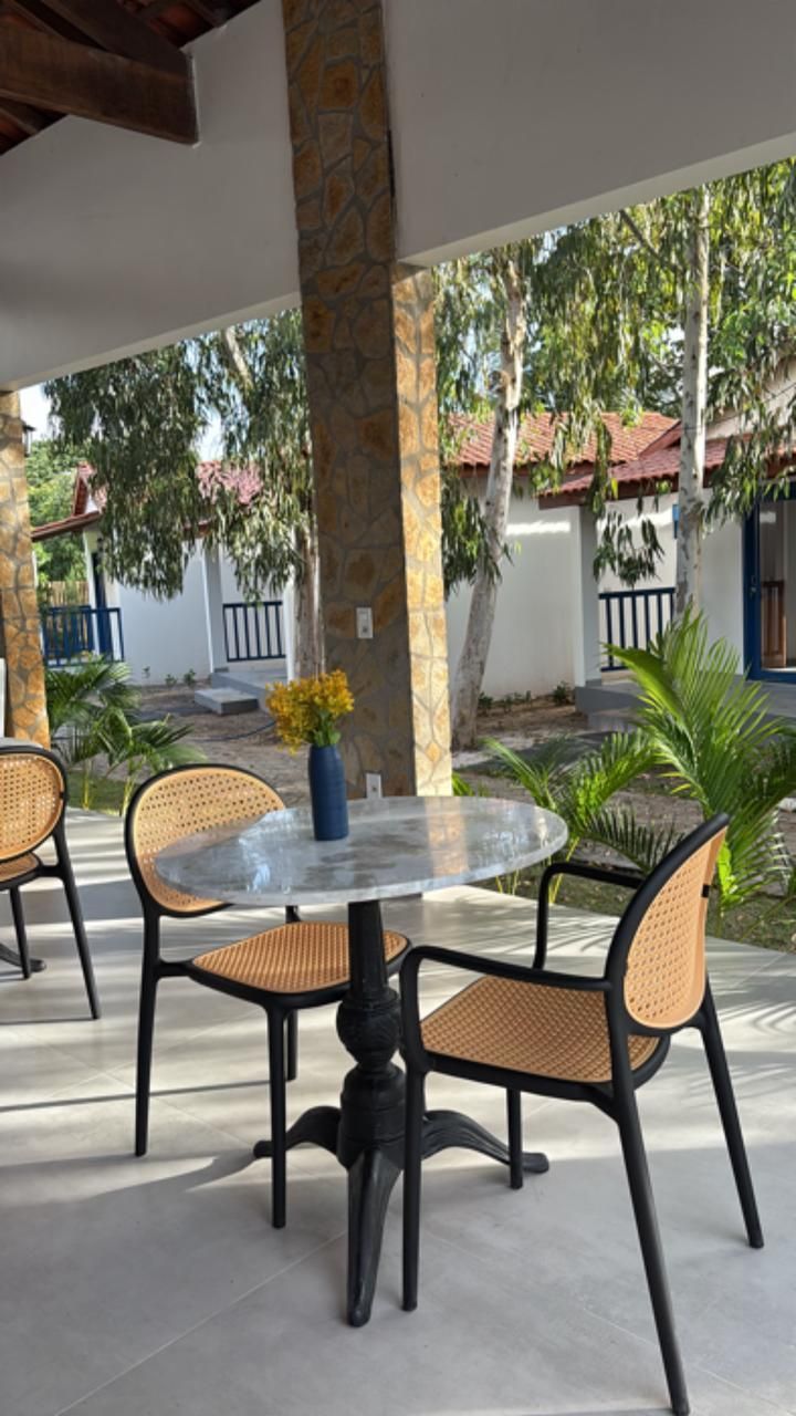 Mesa de café ao ar livre com cadeiras pretas, vaso azul e palmeiras em um pátio sombreado.