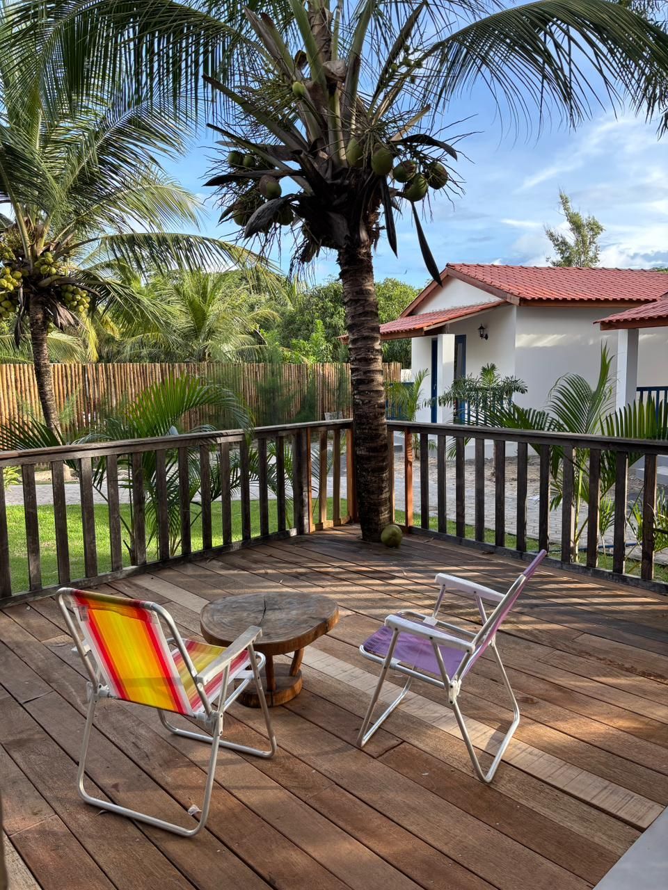 Cadeiras coloridas e uma pequena mesa em um deck de madeira, com vista para palmeiras e uma casa branca com telhado vermelho.
