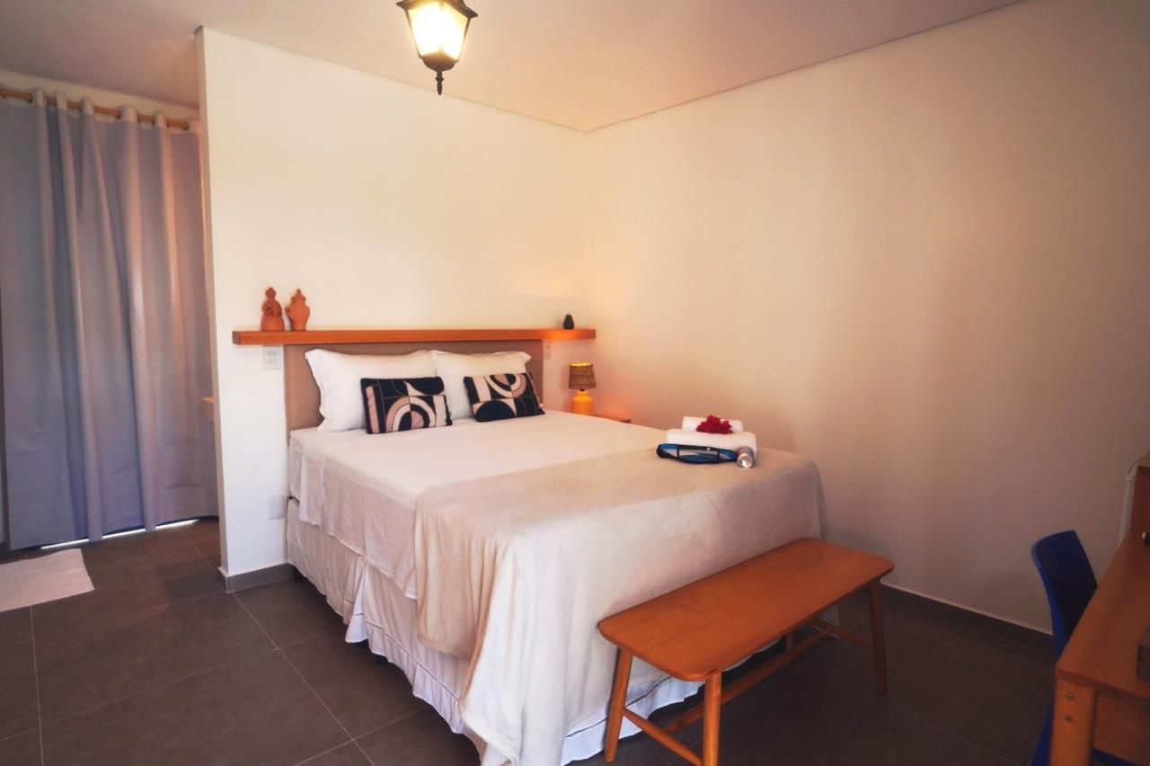 Quarto pequeno com cama arrumada, banco de madeira, escrivaninha e janela com cortina.
