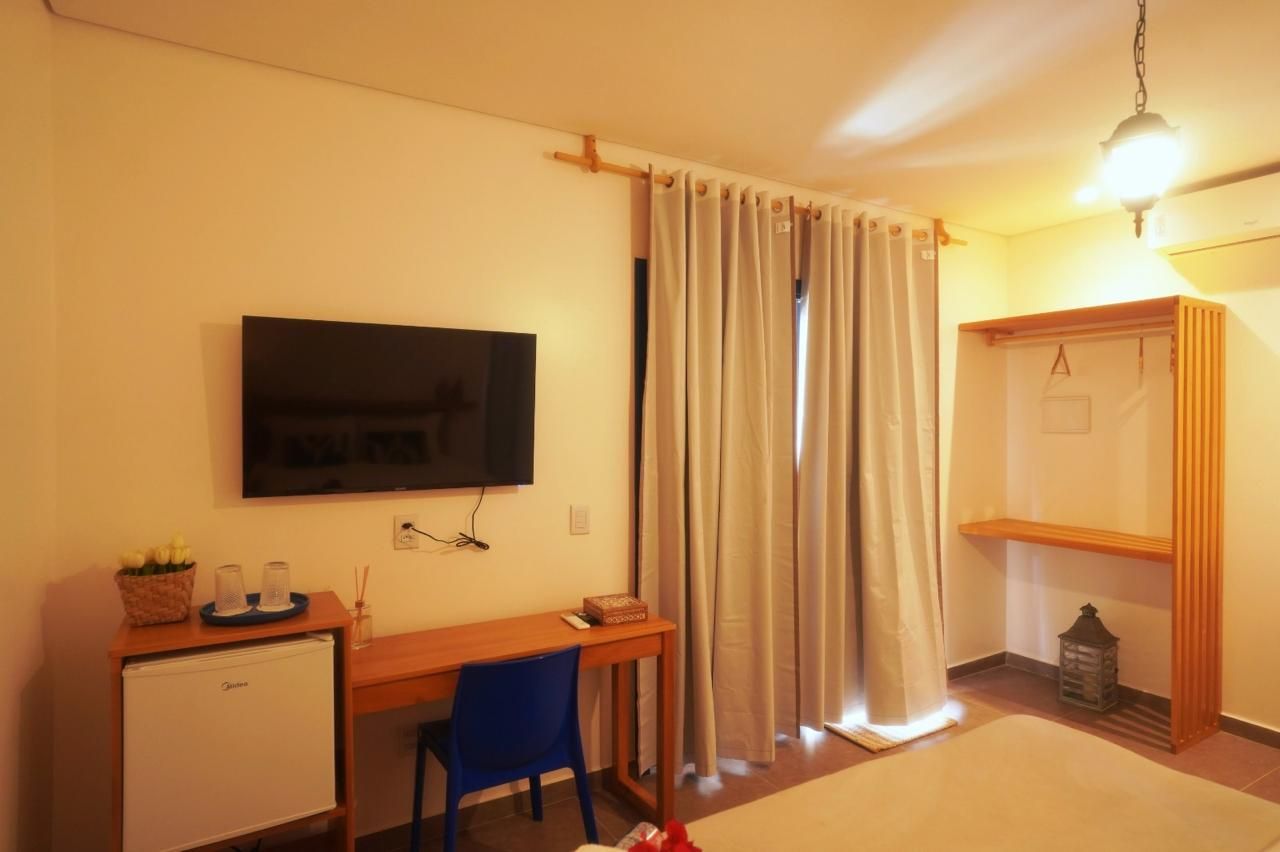 Quarto aconchegante com TV fixada na parede, escrivaninha pequena, cadeira azul, cortinas e um nicho com prateleira de madeira.