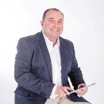 Man in pinstripe blazer smiles, holding a tablet, sitting against white background.
