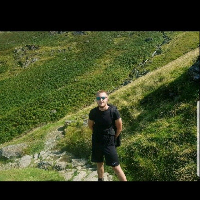 Man in black shirt and shorts standing on a grassy hill, surrounded by green foliage.