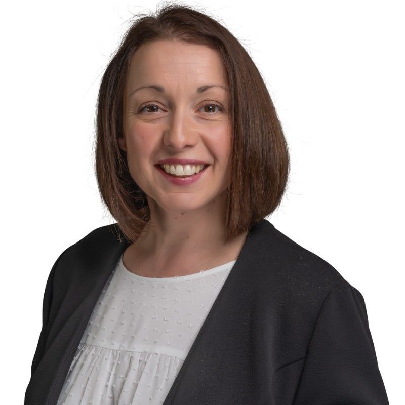 Woman with brown hair, smiling, wearing a white top and black blazer, against a white background.