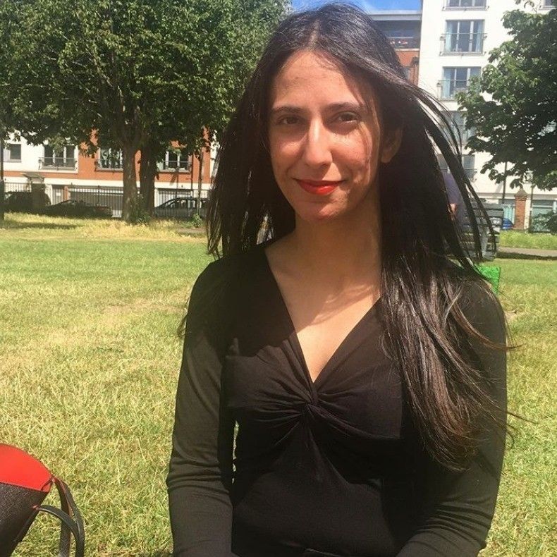 Woman with long dark hair smiles outdoors, wearing a black top. Trees and buildings in the background.