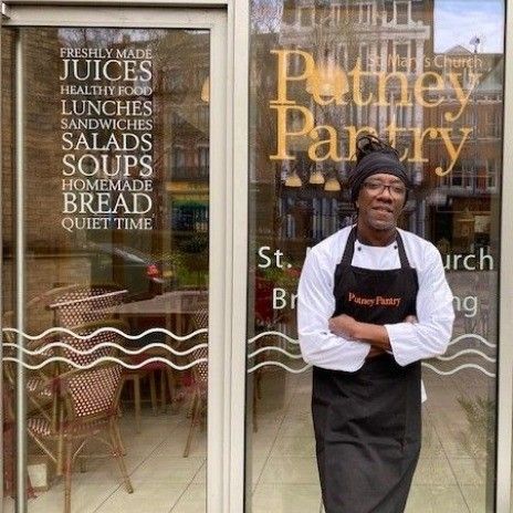 Chef in front of Puthey Pantry cafe. Black apron, white chef coat, menu on window.