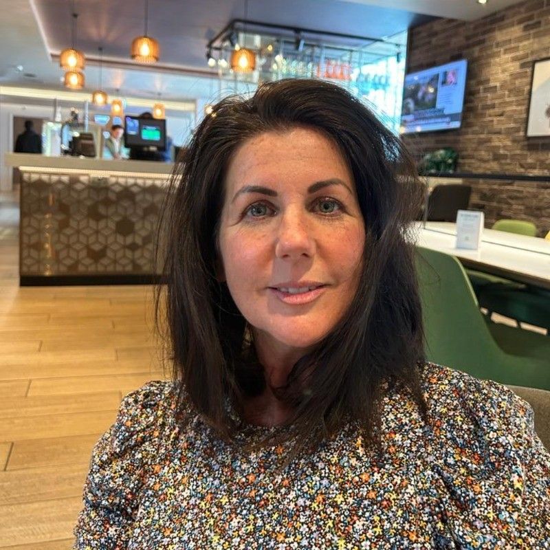 Woman with dark hair smiles in a hotel lobby. Floral blouse, soft lighting, and reception desk in background.