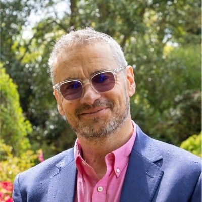 Man wearing sunglasses smiles outdoors; pink shirt, blue blazer, lush greenery background.