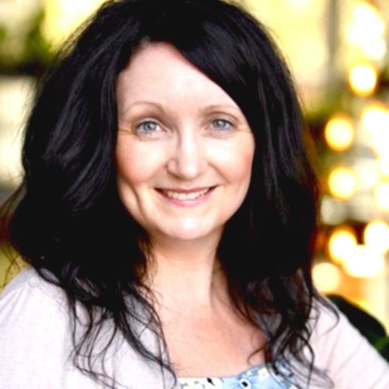 Woman with dark hair smiles, wearing a white cardigan and patterned top, in a bright setting.