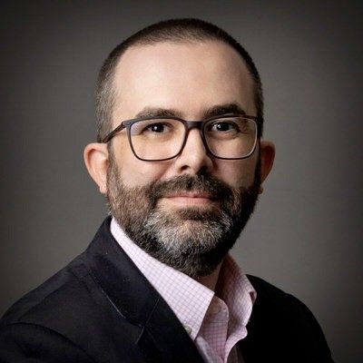 Man with glasses and beard, wearing a blazer and pink shirt, looking at the camera.