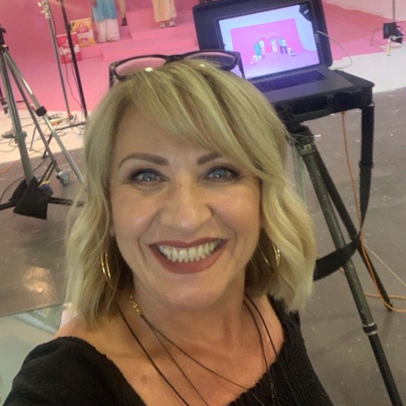 Blonde woman smiling, wearing glasses, in front of a video monitor and filming setup, a pink background.