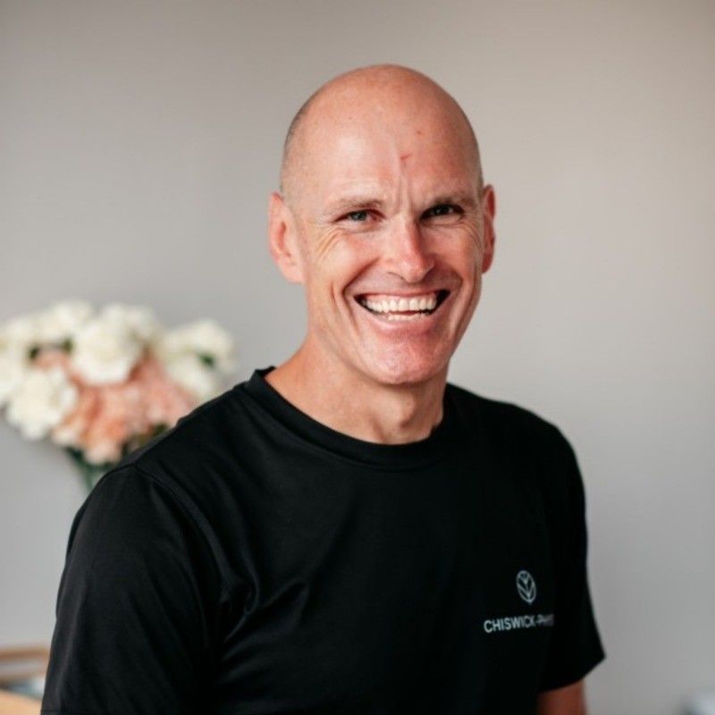 Bald man with a big smile wearing a black shirt, standing in front of flowers in a well-lit room.