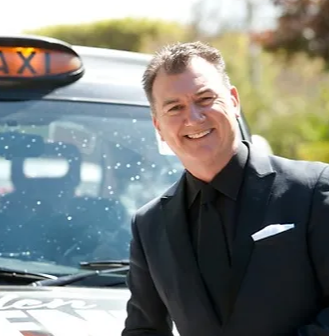 Man in black suit smiles, standing by a London taxi.