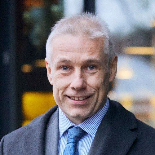 Man in suit smiling, with light blue tie and striped shirt. Gray hair.