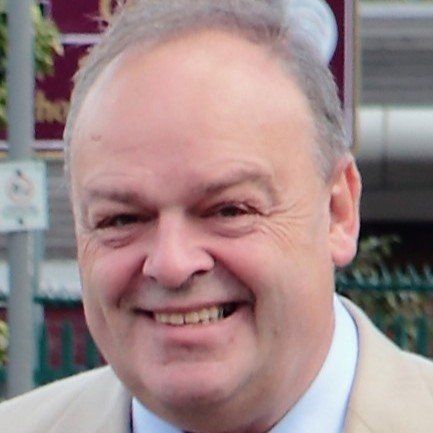 Man smiling outdoors; wearing a light-colored suit jacket.