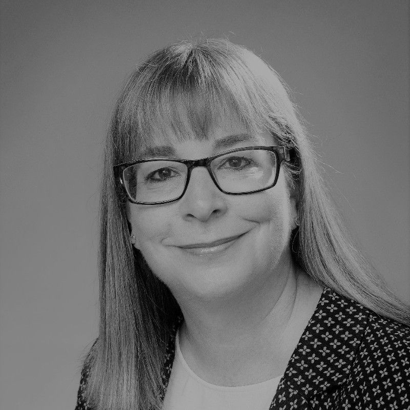 Woman with glasses, smiling, in a patterned blazer, indoors, neutral background.