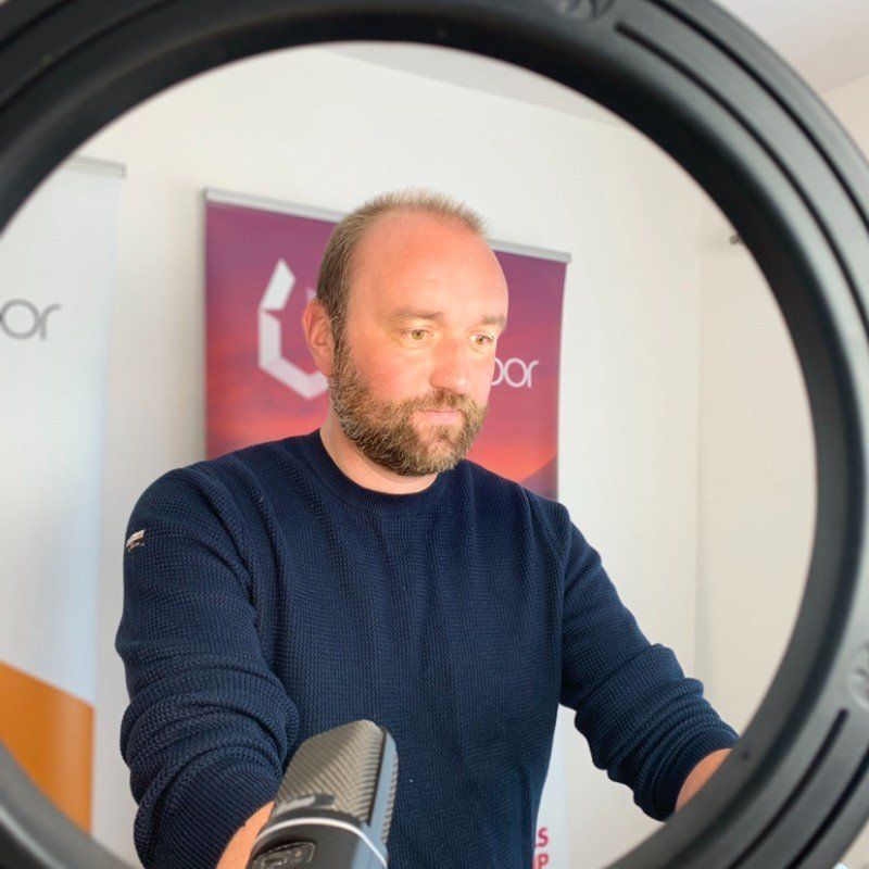 Man with beard in front of ring light and microphone, with banner in the background.