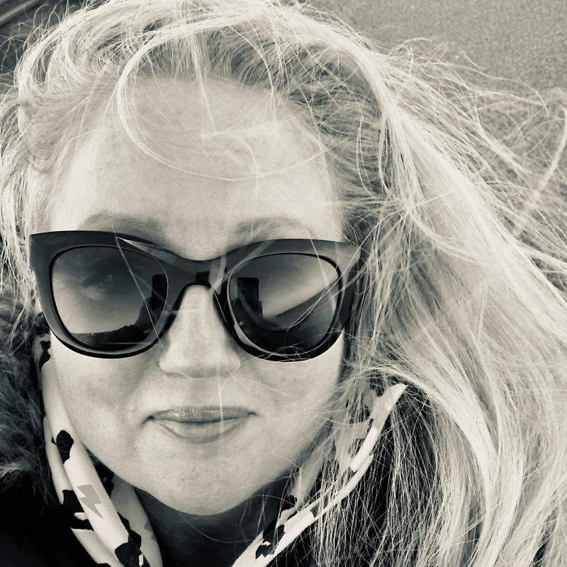 Woman with long, blonde hair wearing large, black sunglasses, and a black and white scarf.