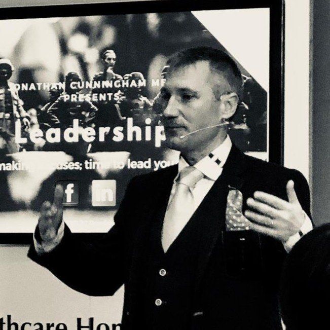 Man giving a presentation, gesturing, wearing a suit. Background includes the word