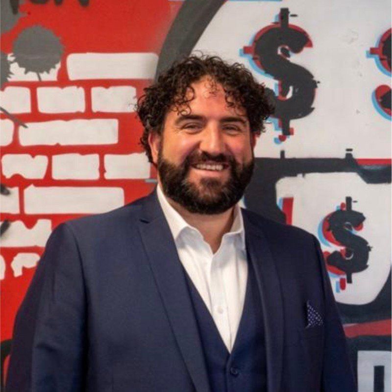 Man in blue suit smiling, standing in front of graffiti with dollar signs.