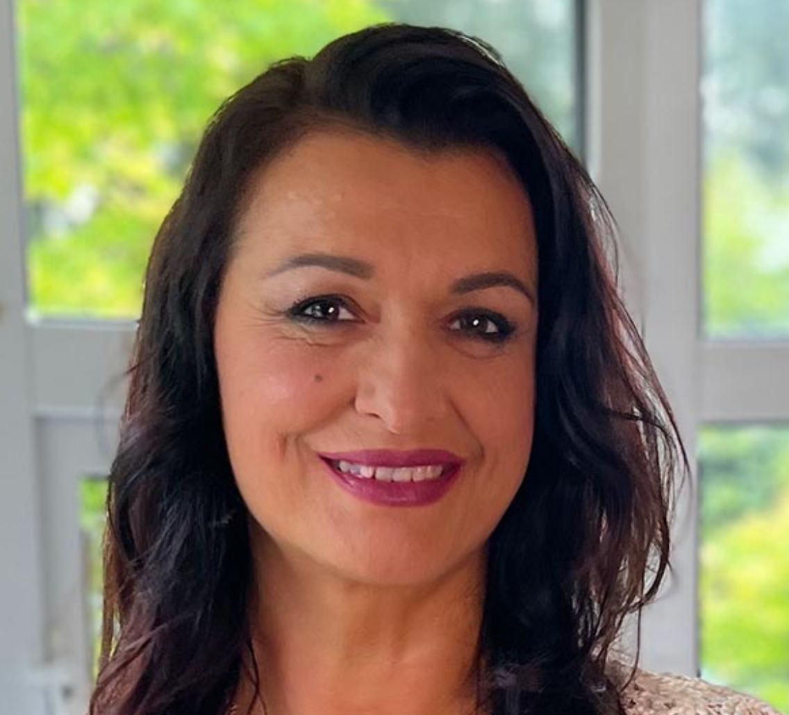 Woman with dark hair, smiling, in front of a window. She is wearing a lace top, and has dark lipstick.
