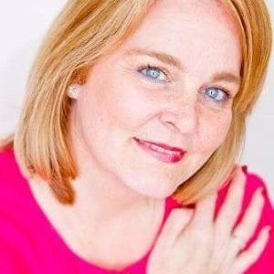 Woman with blonde hair and blue eyes, wearing a pink top, looking at the camera.