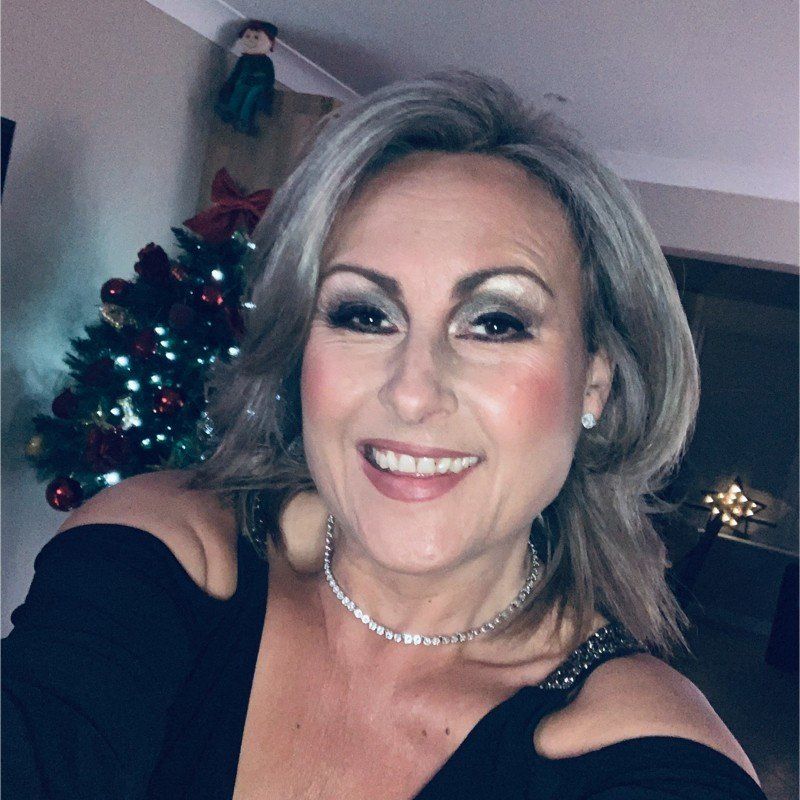 Woman with silver hair smiles, wearing a black top and diamond necklace, by a lit Christmas tree.