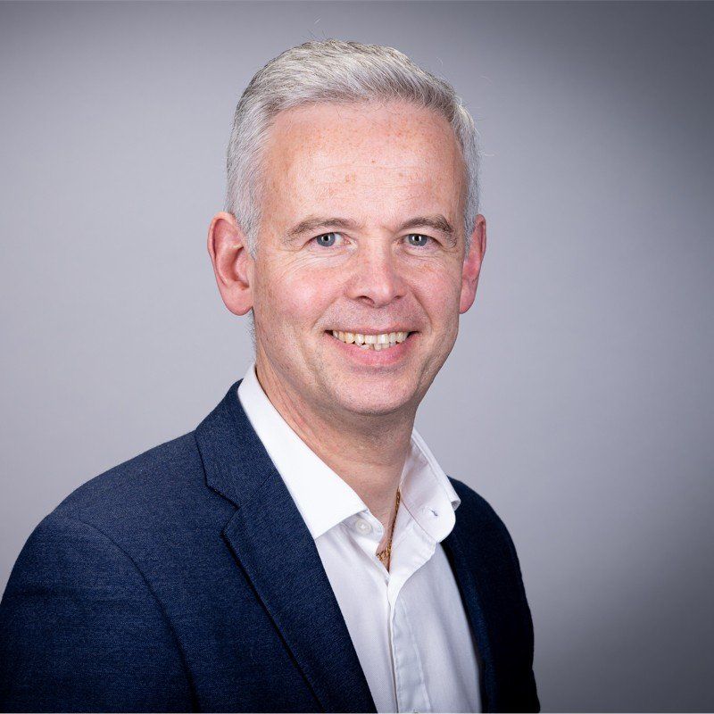 Man with gray hair, smiling, wearing a blue blazer and white shirt, against a gray backdrop.