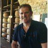 Man smiling in front of shelves holding cheese wheels in a brick-walled room.