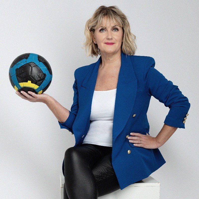 Woman in blue blazer holds a soccer ball. She sits, smiling, in a studio setting.