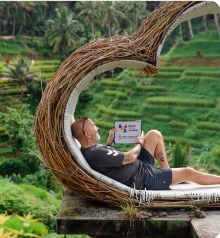 Man reclines in a wicker nest chair, reading a tablet overlooking rice terraces in Bali.