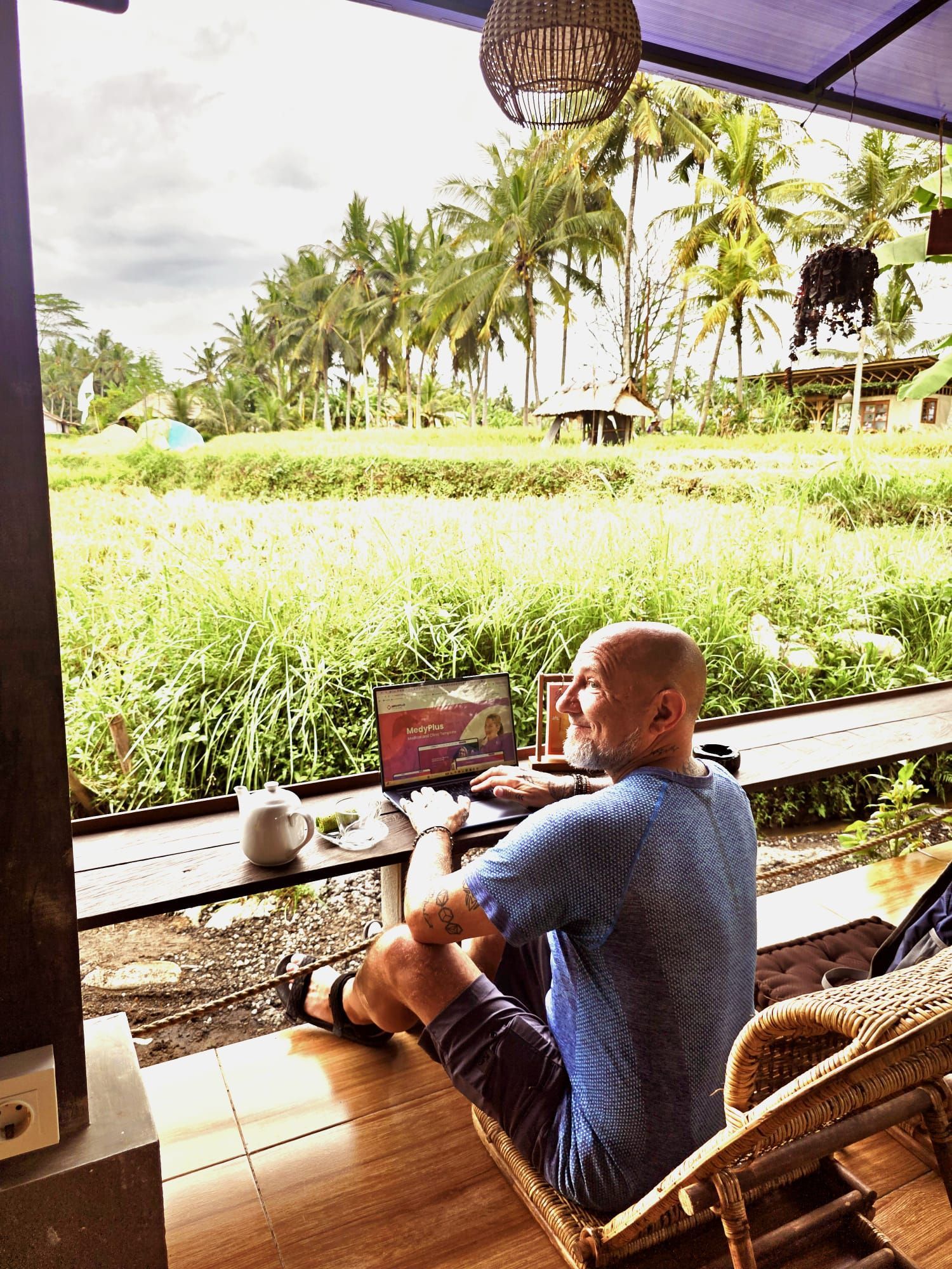Man working on a laptop, seated on a porch overlooking a green field with palm trees.