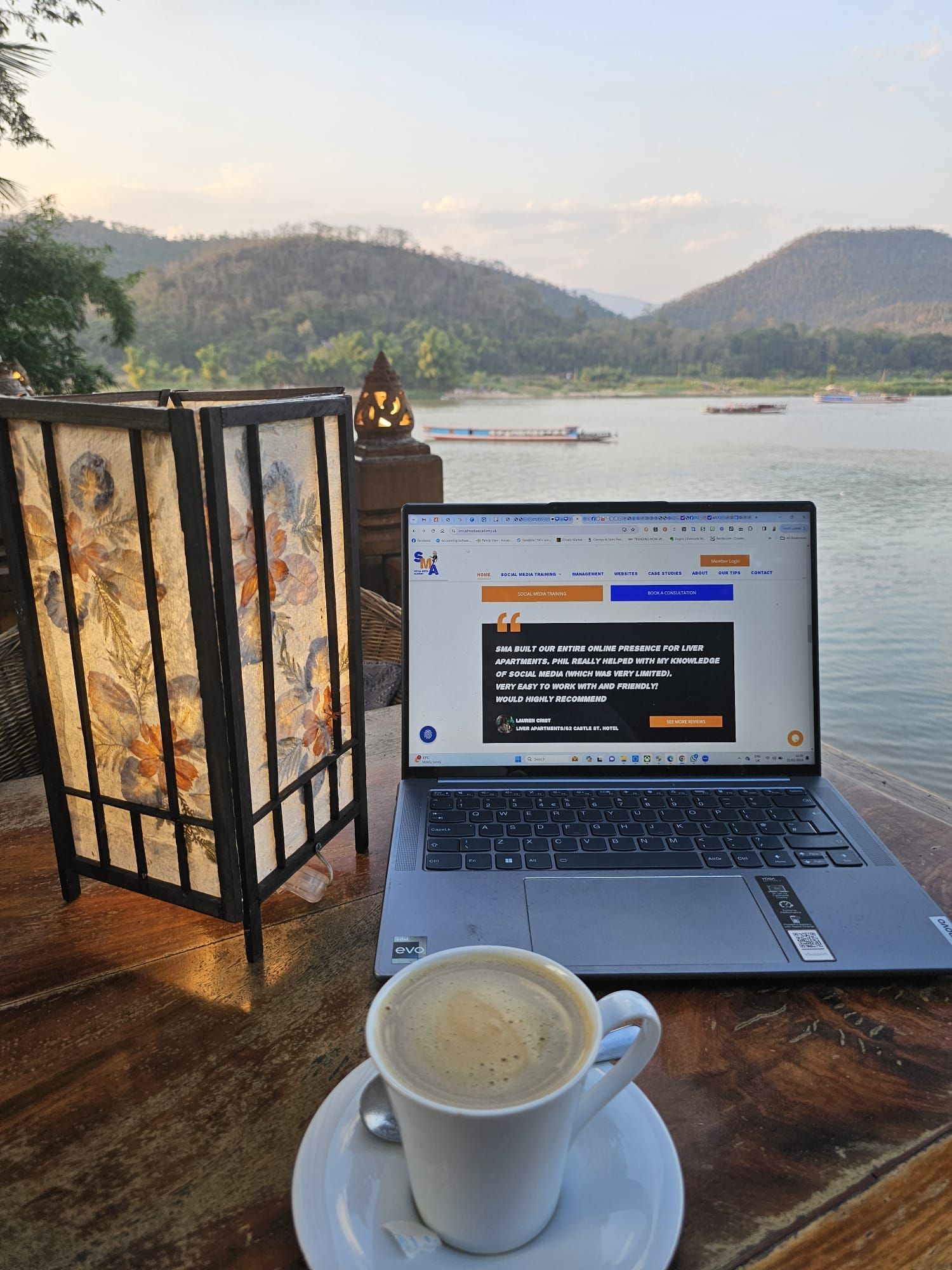Laptop with coffee on a wooden table, lit lamp, river and mountains in background.