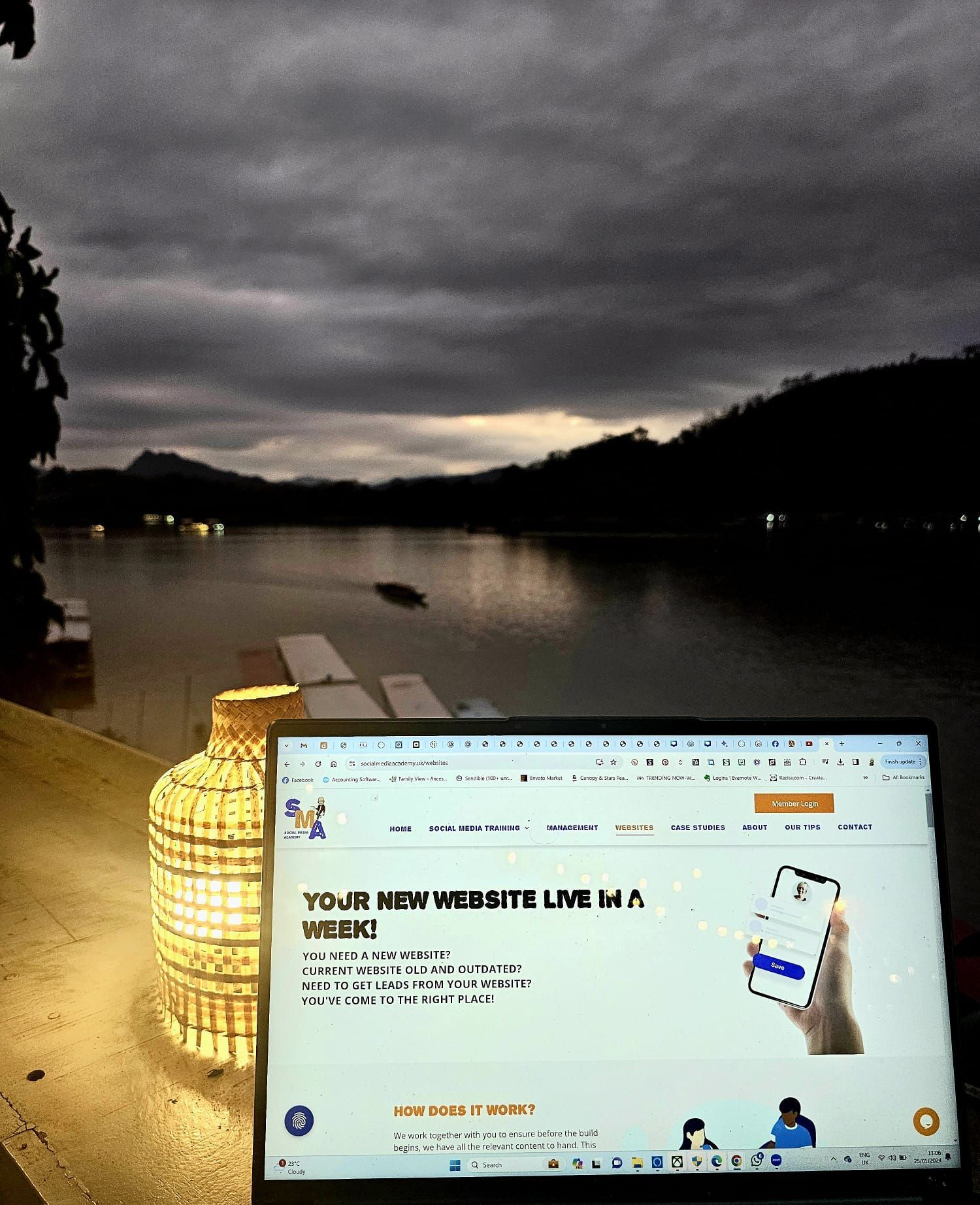 Laptop displaying website, lit by a lamp, overlooking a dark lake and mountains at dusk.
