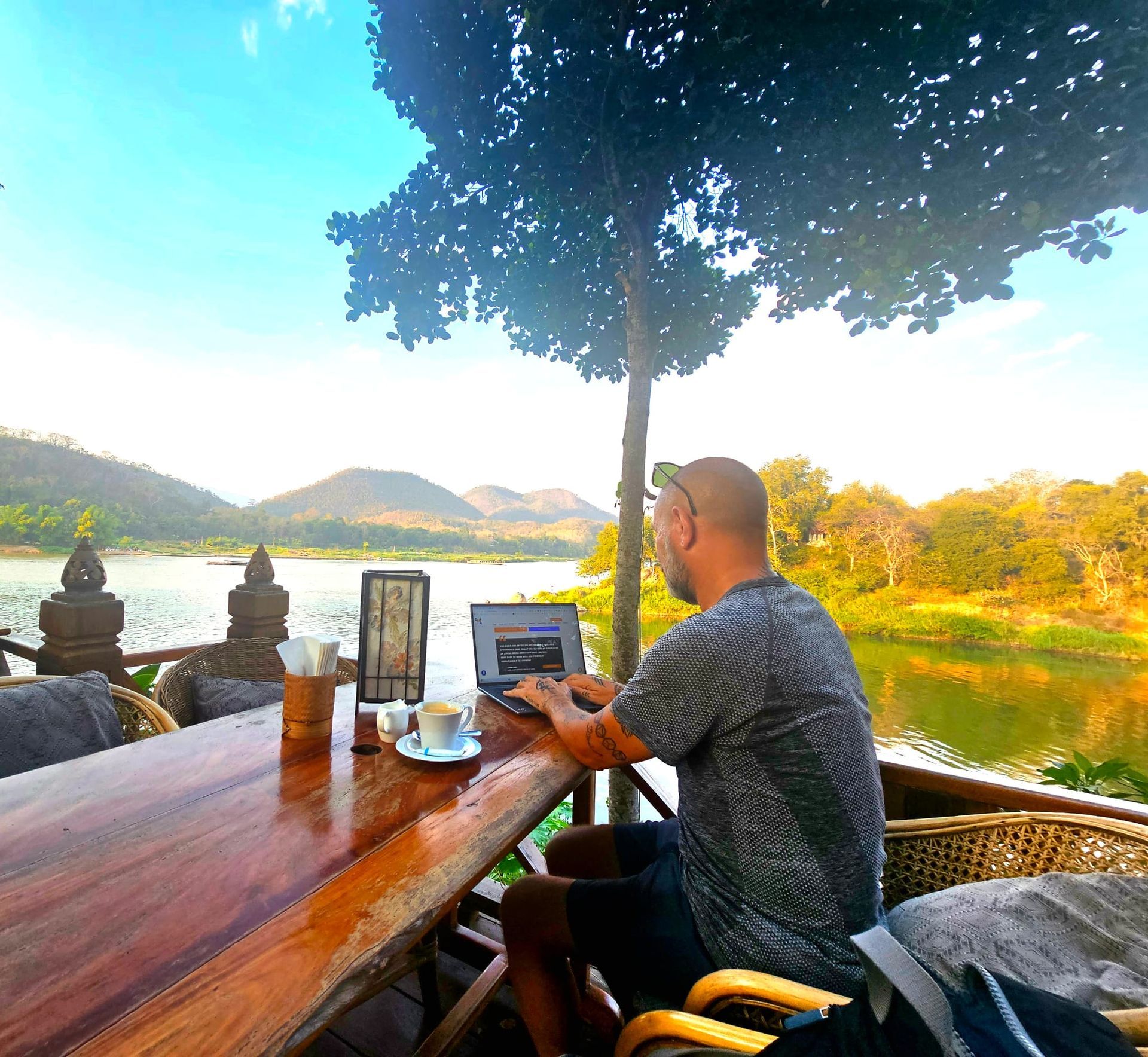 Man working on a laptop at a waterside cafe. Overlooks lake and mountains on a sunny day.