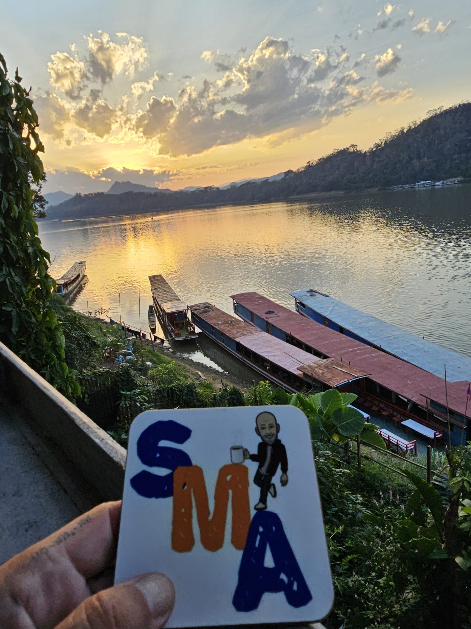 Hand holding SMA coaster with river and sunset view.
