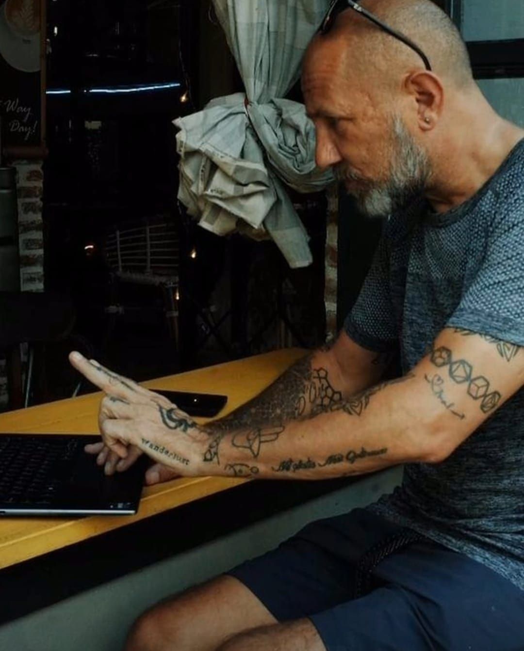 Man with arm tattoos pointing at a laptop on a yellow surface, indoors.