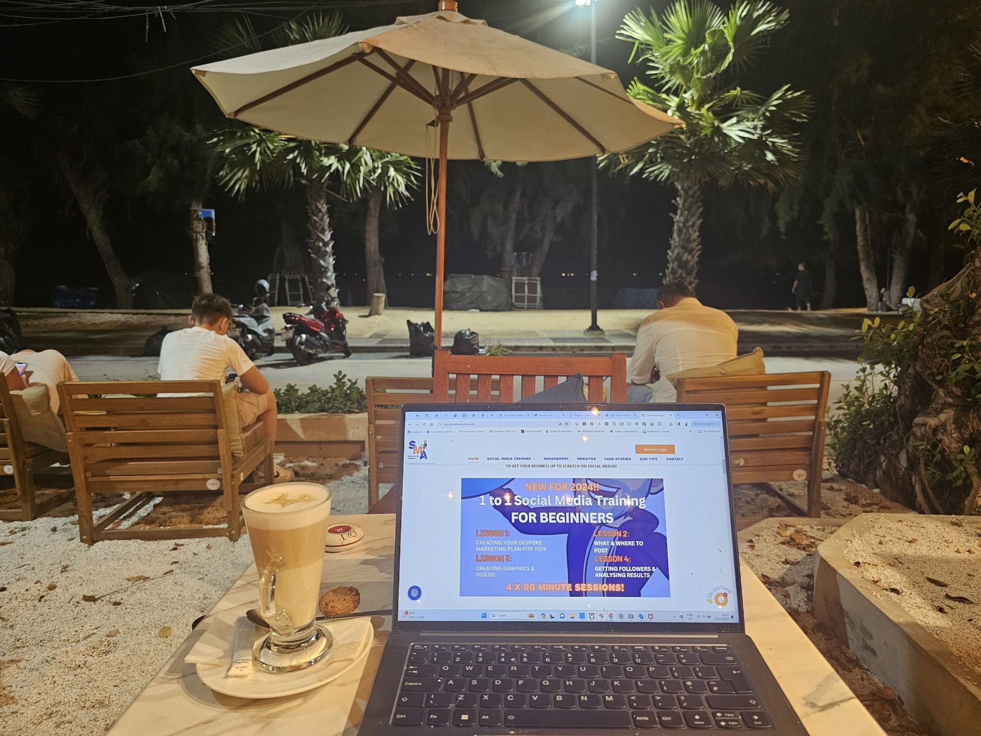 Laptop with website open, coffee, and outdoor cafe setting at night with two people.