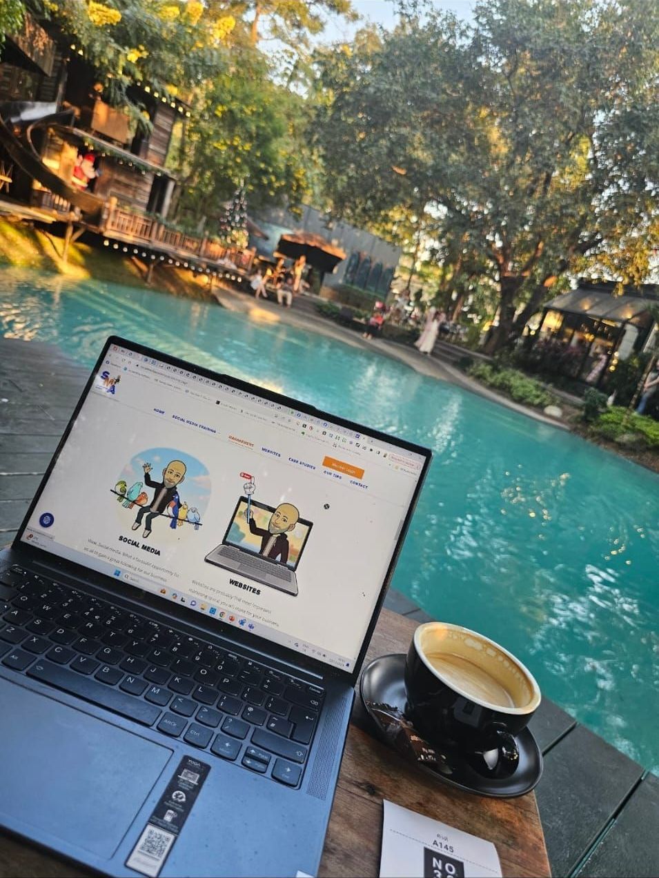 Laptop with website open next to coffee cup and pool. Outdoor setting with trees and wooden structures.